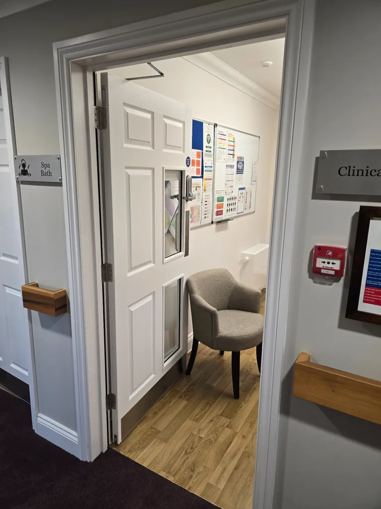Open white door with vision panels leading into clinic room with chair, wall notice board and wooden flooring